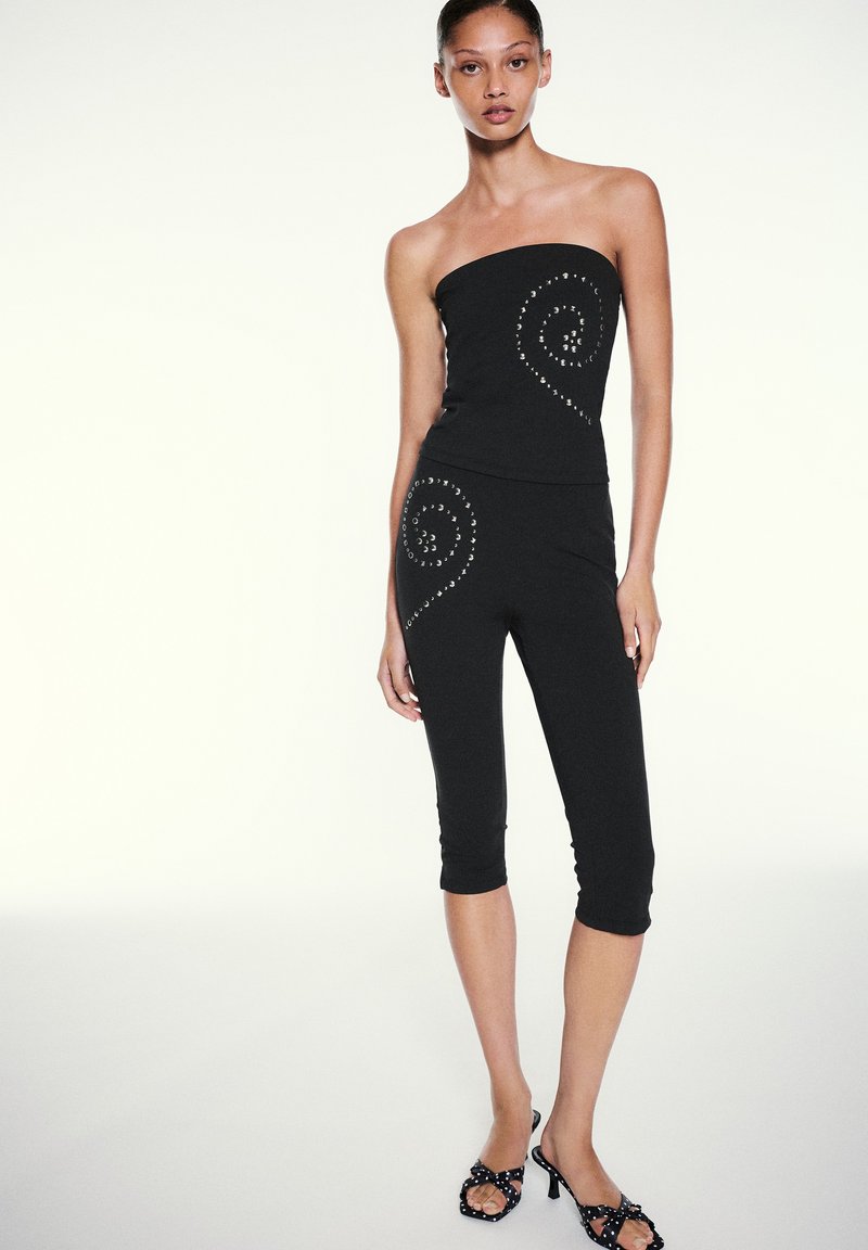 Femme portant un haut bustier noir et un pantalon court avec des motifs de clous en spirale, associés à des sandales noires à talons hauts sur un fond clair uni.