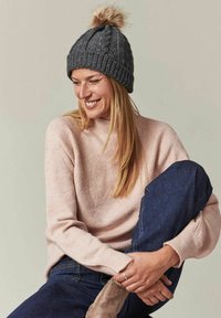 Bonnet en tricot gris avec un pompon en fourrure, porté avec un sweat-shirt rose clair affichant un motif texturé subtil, associé à un jean bleu foncé.