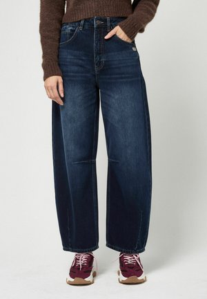 Persoon draagt donkerblauwe jeans met wijde pijpen, een bruine trui en kastanjebruine sneakers met witte zolen en lichtroze veters, staand tegen een effen achtergrond.