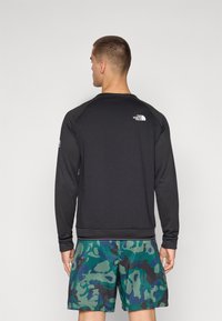 Zwarte langemouwige shirt van textuurstof met ribbelpatroon, voorzien van een klein wit logo op de bovenrug, gecombineerd met groene camouflage shorts.
