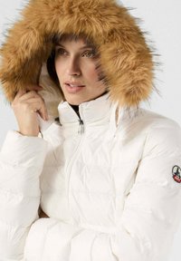 Chaqueta acolchada blanca con aislamiento y capucha de piel sintética marrón. Presenta un cuello alto y cierre con cremallera. Parche de logo en la manga izquierda.