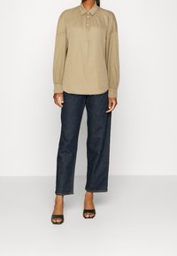 Chemise beige à manches longues, boutonnée, avec détail froncé, portée avec un jean large bleu foncé et des sandales à talons ouverts noires.