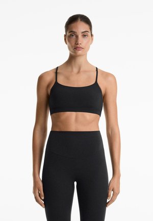 Schwarzer Sport-BH mit dünnen Trägern und quadratischem Ausschnitt, kombiniert mit hochtaillierten schwarzen Leggings. Der Stoff wirkt glatt und dehnbar.