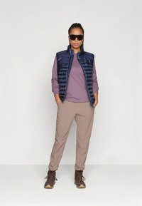 Tamsiai mėlyna pūsta liemenė virš purpurinės ilgomis rankovėmis palaidinės, derinama su smėlio spalvos kelnėmis ir rudais žygių batais, dėvima su tamsiais akiniais nuo saulės.