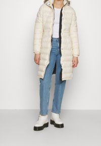 Długi, puszysty płaszcz w kolorze kremowym z kapturem, z zamkiem błyskawicznym i zapięciem na napy. Połączony z niebieskimi dżinsami i białymi botkami sięgającymi do kostek.