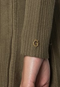 Manica di un maglione lavorato a maglia verde oliva con texture a coste, che presenta un accento in oro con la lettera 'G' vicino al polsino.