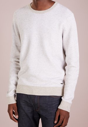 Hombre vistiendo un suéter de punto texturizado color beige claro con puños y dobladillo acanalados, combinado con jeans azul oscuro.