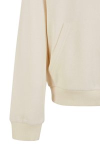 Beiger Sweatshirtärmel mit geripptem Bündchen und vorderer Kängurutasche, die die Stoffstruktur und Nähte zeigt.