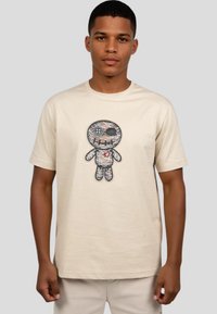 Beige cotton t-shirt featuring a printed graphic of a stitched character with black accents and a red heart detail on its chest.