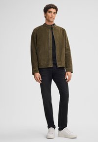 Olijfgroene suedebroek met een opstaande kraag, ritssluiting aan de voorkant en twee zijzakken, gecombineerd met zwarte jeans en witte sneakers.