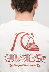 Άτομο που φοράει λευκό πουκάμισο με κόκκινο στυλιζαρισμένο κύμα και λογότυπο βουνού και κείμενο "Quiksilver The Original Boardshort Co." στην πλάτη.