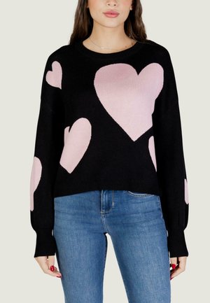 Frau trägt einen schwarzen Pullover mit großen pinkfarbenen Herzen und blaue Jeans, gezeigt von Hals bis Hüfte vor einem einfarbigen Hintergrund.