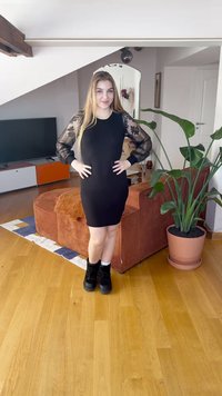 Vestido justo preto com mangas compridas em renda floral transparente, usado com botas pretas até ao tornozelo. O fundo apresenta uma planta e um sofá elegante.