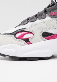 Gros plan sur une sneaker en mesh blanc, en suède gris, avec des accents noirs et roses, et une épaisse semelle texturée blanche sur un fond uni.