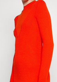 Robe en tricot ribbed dans un rouge-orange vibrant, avec des manches longues et une silhouette ajustée. L'encolure est de coupe arrondie haute avec un détail angulaire.