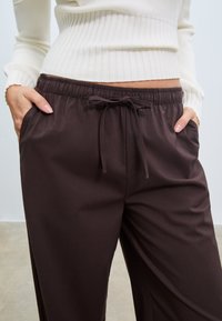 Braune, weit geschnittene Hosen mit Kordelzug und Seitentaschen, aus einem glatten Stoff mit weicher Textur und leichtem Glanz.