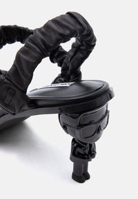 Μαύρο σανδάλι με ραμμένες λουρίδες και μικρό τακούνι σε σχήμα φιγούρας που φορά γυαλιά ηλίου και κοστούμι, σε λευκό φόντο.