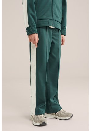 Osoba ubrana w zielone spodnie dresowe z białymi paskami po bokach, w beżowo-szare sneakersy, stojąca na jasnym tle.