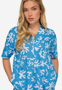 Top de manga corta, azul con un patrón blanco parecido al coral. Cuenta con un escote en V y bolsillos, hecho de una tela ligera.