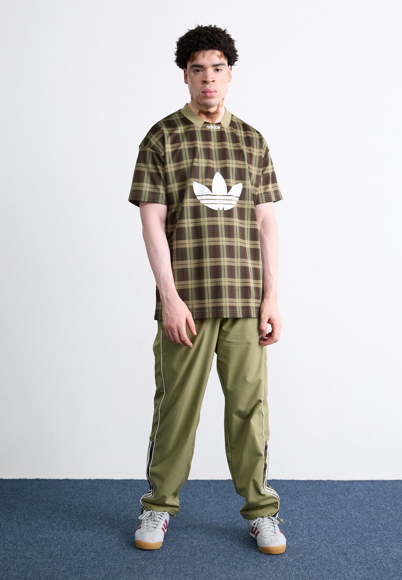 Zielona kratkowana koszula z krótkim rękawem z białym logo, w zestawieniu z oliwkowymi spodniami z bocznymi paskami oraz szarymi sneakami z czerwonymi akcentami.