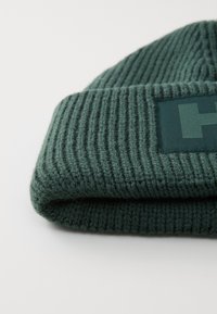 Grønn strikket lue med ribbestruktur, med en rektangulær stofflapp med logo. Mykt, fleksibelt materiale, med rullet kantdesign.