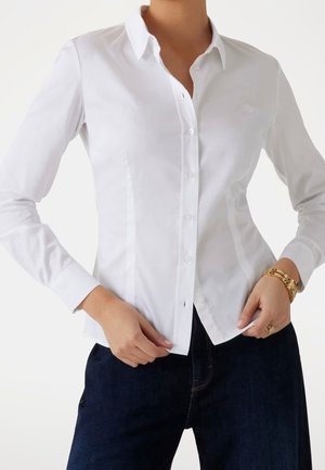 Femme portant une chemise blanche ajustée à boutons avec manches longues et un pantalon bleu foncé, accessoirisée d'une montre en or et d'une bague.