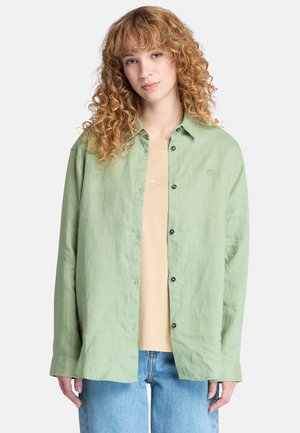 Light green long-sleeve shirt with a button-down front, featuring a soft, textured fabric and a small logo on the chest. Paired with a beige top.