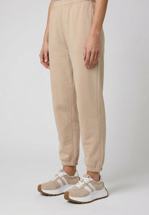 Person iført beige joggebukser og matchende beige og hvide sneakers, stående mod en ensfarvet lys baggrund.