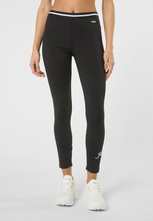 Kobieta ubrana w czarne legginsy FILA z białym logo i paskiem w paski, w parze z białymi sneakersami FILA, stojąca na jednolitym tle.