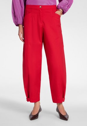 Frau trägt leuchtend rote, hoch taillierte Hosen mit Knopfmanschetten, kombiniert mit einer lila Langarmbluse und dunklen, spitzen Absätzen.