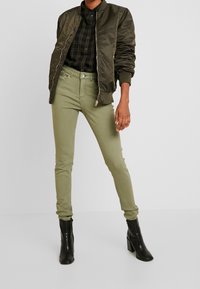 Veste bomber vert olive avec fermeture éclair, associée à un jean skinny vert olive et des bottes à talons noires. Chemise à carreaux sous la veste.