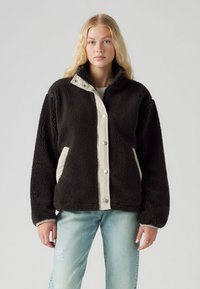 Chaqueta de forro polar negra con tapeta de botones beige y bolsillos laterales; cuenta con un cuello alto, textura suave y un ajuste relajado.