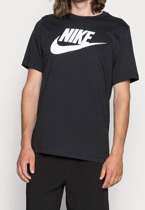 Personne aux cheveux longs portant un t-shirt noir Nike avec un grand logo blanc sur la poitrine, debout, les bras détendus.