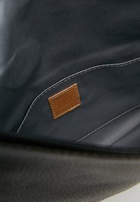 Wnętrze czarnej torebki z gładką podszewką, z widoczną etykietą z brązowej skóry i białym przeszyciem. Brak widocznych dodatkowych kieszeni.