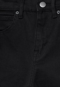 Gros plan sur un jean en denim noir montrant la ceinture, les passants de ceinture, le bouton en métal, les rivets et les détails des coutures de la poche avant.