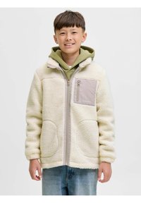 Kurtka polarowa w kremowym kolorze o miękkiej fakturze, z zamkiem z przodu i beżową kieszenią. Noszona na zielonym hoodie, w komplecie z niebieskimi dżinsami.