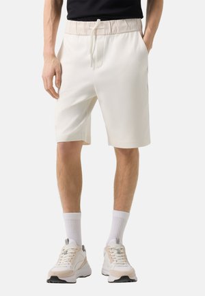 Mann trägt weiße Shorts mit Kordelzug, weiße Sportsocken und weiße Turnschuhe vor neutralem Hintergrund.