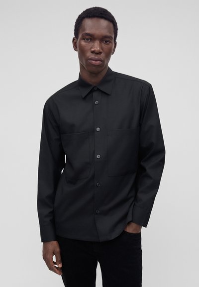 Homme portant une chemise noire à manches longues boutonnée avec deux poches poitrine, debout devant un fond gris uni.