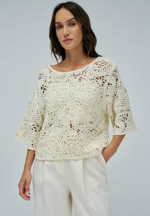 Haut en crochet crème avec une coupe loose, doté de motifs floraux délicats et de manches longueur coude. Assorti à un pantalon crème pour un look coordonné.