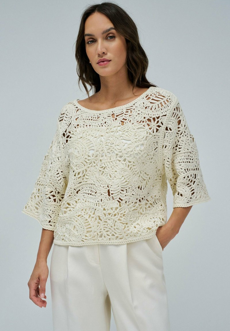 Cremfarbener Häkeloberteil mit lockerer Passform, das aufwändige florale Muster und dreiviertellange Ärmel aufweist. Kombiniert mit cremfarbenen Hosen für einen stimmigen Look.