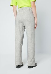 En person som bär ljusgrå lösa byxor, en neongrön t-shirt med kort ärm och svarta skor, står mot en enfärgad ljus bakgrund.