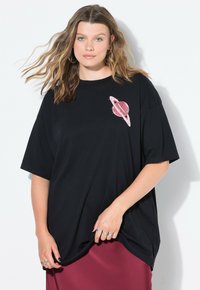 Zwarte oversized t-shirt van katoen, voorzien van een roze planeetgraphic met ringen op de linkerkant van de borst. Soepele textuur, ronde hals.