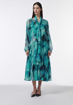 Vrouw draagt een lange, luchtige teal en zwarte jurk met een patroon, doorschijnende lange mouwen en een strik bij de hals, gecombineerd met zwarte puntige hakken.