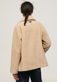 Osoba z warkoczem nosi beżową kurtkę z polarowego materiału i ciemnoniebieskie dżinsy, pokazana od tyłu na jednolitym jasnym tle.