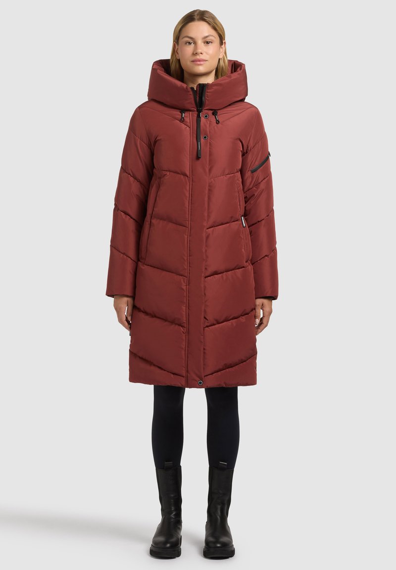 Piumino rosso con collo alto e cappuccio, presenta una cerniera frontale e tasche laterali. Il tessuto ha una texture liscia ed è imbottito.