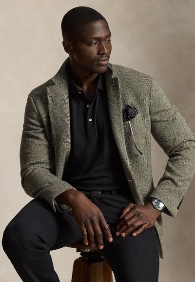 Mann trägt einen Fischgräten-Blazer über einem schwarzen Poloshirt, sitzt auf einem Holzschemel und schaut leicht nach links.