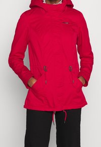 Personne portant une veste à capuche rouge vif avec des poches avant et des cordons de serrage, associée à un pantalon noir, les mains dans les poches.