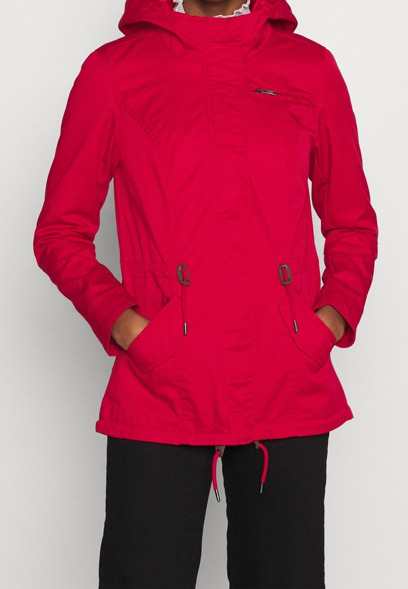 Personne portant une veste à capuche rouge vif avec des poches avant et des cordons de serrage, associée à un pantalon noir, les mains dans les poches.