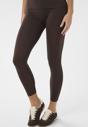 Persona che indossa leggings aderenti marrone scuro e scarpe da ginnastica marrone scuro con lacci e suole bianche, in piedi su uno sfondo bianco.