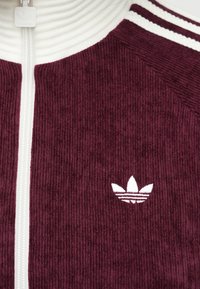Burgundijas kordūras jaka ar rāvējslēdzēju, ar baltu ribotu apkakli, kas rotāta ar mazu baltu logotipu kreisajā pusē un svītrainiem akcentiem plecos.